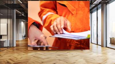 A worker in orange coverall uniform is opening documents to make a discussion on working procedure, safety audit in operation action photo. Close-up and selective focus on human's hand. Wall mural