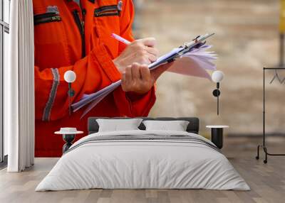 Action of a safety officer in full PPE coverall is writing note on paper document during perform safety audit at construction worksite. Industrial expert working scene. Selective focus. Wall mural