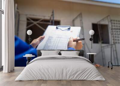 An architect is checking on house building quality checklist form to inspecting the progress with building house as blurred background. Construction industrial working scene, close-up. Wall mural