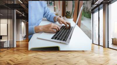 A happy, creative businesswoman sits at a wooden desk in a cozy cafe, using a laptop and smartphone for online communication. She types on the keyboard, reflecting a modern, connected lifestyle. Wall mural