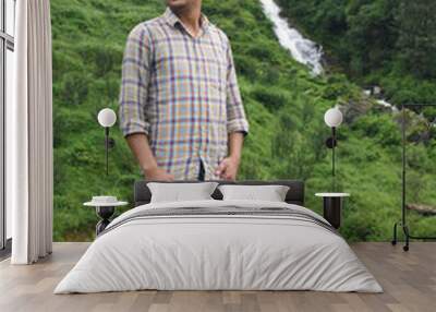 Male model standing outdoors left hand in pocket and looking away. Waterfall and lush greeenery in background Wall mural