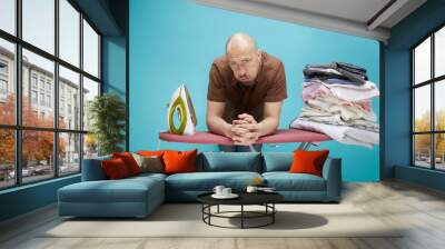 A funny bald man with emotions using electrical iron on ironing board over on blue background. Wall mural