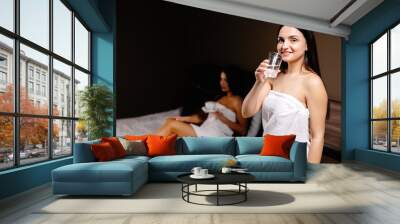 Cute young woman holding a glass of water in the morning while standing in the bedroom and behind her is her friend lying on the bed and drinking coffee Wall mural