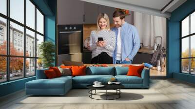 Smiling loving couple cooking salad together while standing on a kitchen at home and using mobile phone. Cute couple man and woman taking photo of food and taking pictures together while cooking. Wall mural