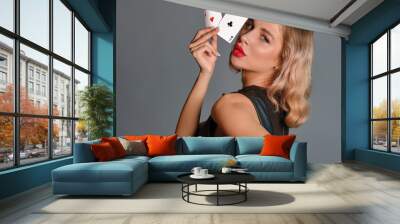 Blonde girl in black dress showing two playing cards, posing sideways against gray background. Gambling entertainment, poker, casino. Close-up. Wall mural