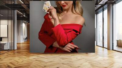 Blonde model in red dress and black earrings. She smiling, showing two playing cards, posing on gray studio background. Poker, casino. Close-up Wall mural