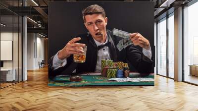 Handsome emotional man is playing poker sitting at the table in casino against a white spotlight. Wall mural