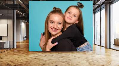 Mom and daughter with a funny hairstyles, dressed in black shirts and blue denim jeans are posing against a blue studio background. Close-up shot. Wall mural