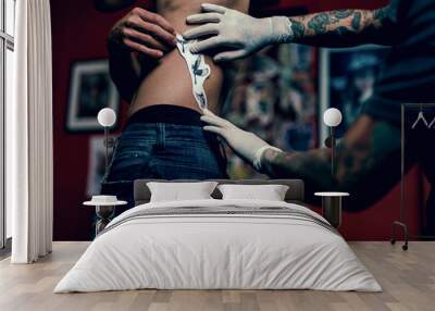 Close-up hands of master Tattoo artist in white gloves sticking paper with pattern on client hand on a Man waist, in a modern in Studio lowlight. Wall mural