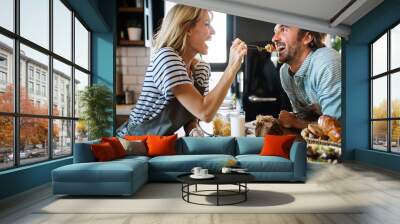 Beautiful young couple is talking and smiling while cooking healthy food in kitchen at home Wall mural