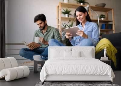 Young couple reading together book and ebook, sitting on couch Wall mural