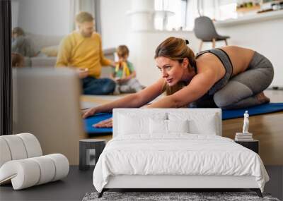 Young woman, mother exercising at home in living room, father playing with kids in background. Wall mural