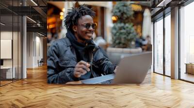 A cheerful young African American man with dreadlocks, wearing sunglasses and a black jacket, is using a laptop outdoors at a cafe. Wall mural