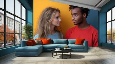 Two people stand against a solid yellow and blue background, gazing at each other with curiosity. Their expressions convey a sense of connection. Wall mural