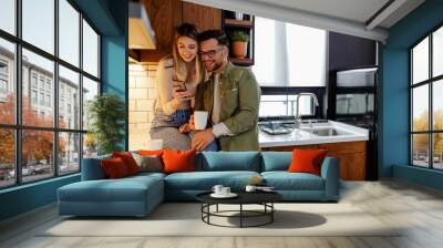 Beautiful young couple is using a phone and smiling while drinking a coffee in kitchen at home. Wall mural