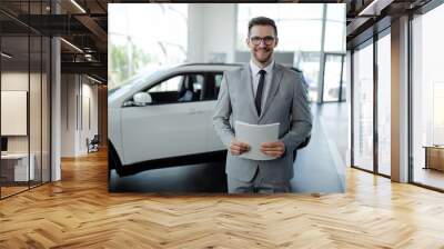 Confident young car dealer standing in showroom and holding a paper while posing. Wall mural