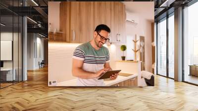 Handsome young man working on a tablet computer while standing in his modern kitchen Wall mural