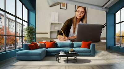 Pensive female administrative assistant checking report on laptop reading information, noting data. Wall mural