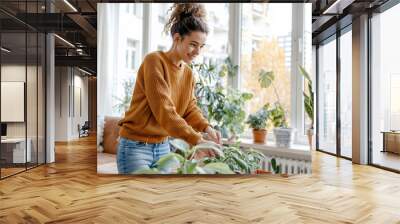 A young woman in a yellow sweater tends to her indoor plants in a bright and modern apartment. Wall mural