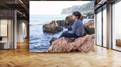 The boy is sitting on a rock by the sea. Wall mural