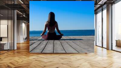 slender fit girl demonstrates Sukhasana yoga meditation pose at wooden flooring against blue background of sky and sea horizon in summer at sunset   Wall mural