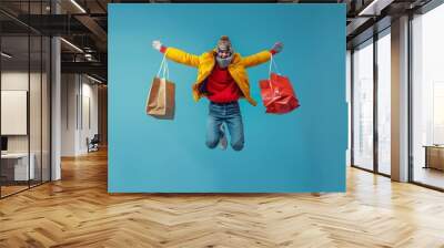 Excited man in colorful clothes leaping with shopping bags in hand, captured mid-air against a clear blue sky. Wall mural