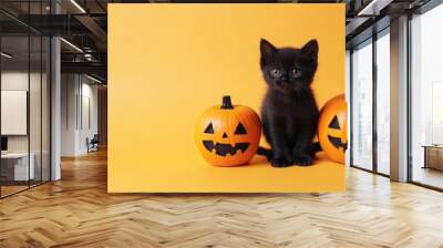 Black kitten between two carved pumpkins, orange background Wall mural