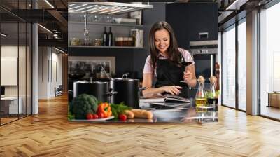 Cheerful young woman in apron on modern kitchen with cookbook an Wall mural