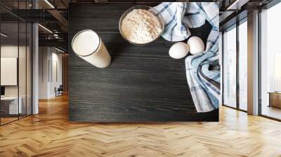 The process of preparation in detail. Cooking, kneading dough. A glass of milk, 2 eggs, a saucer with flour and a blue napkin on a black wooden table Wall mural