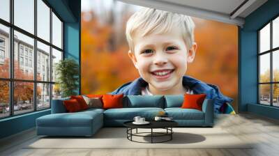 A young Caucasian boy with short blonde hair wearing a colorful striped jacket, smiling joyfully in an outdoor setting with blurred autumn foliage in the background Wall mural