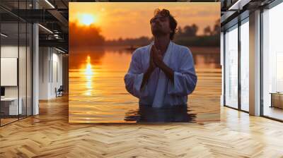 A young man wearing a white robe, standing in the river water after the baptism, praying with clasped hands and closed eyes, looking up to the sky. Sunset time. Male Christian person religious ritual Wall mural