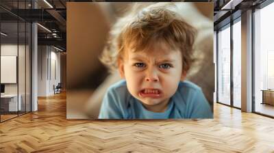 Closeup of the unhappy little male toddler boy, baby in the bad mood sitting on the sofa in a living room, looking at the camera with nervous and frustrated face expression, displeased weeping infant Wall mural