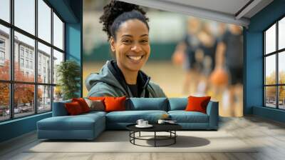 Portrait of a happy African American basketball coach, pretty woman standing on the hardwood court in the basketball gym interior, looking at the camera and smiling. Players blurred in the background Wall mural