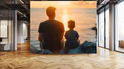 Rearview photography of a father and son sitting together on a rock or cliff on the sea coast, wearing black t shirts and looking at the ocean waves during the golden hour sunset sky, vacation Wall mural