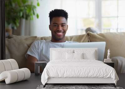 Young African American man sitting on a sofa or couch in the living room at a house or home, and using a laptop computer device. Male adult freelancing, smiling at the camera and holding a notebook Wall mural