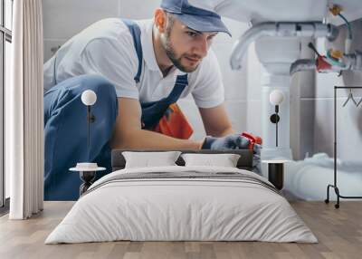 Young handsome plumber wearing a working uniform and a cap, kneeling down in the bathroom to fix or repair the white pipes under the sink. Home service or maintenance, professional handyman job Wall mural