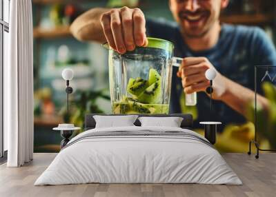 Young man smiling in the background, placing a hand on the electric blender mixer device, standing in the kitchen, making healthy nutritious green smoothie with tropical and exotic kiwi fruit Wall mural