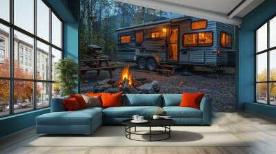 A large camping trailer is parked beside a crackling campfire in an evening forest setting. The soft light from the trailer peers out into the tranquil exterior, creating a cozy ambiance. Wall mural