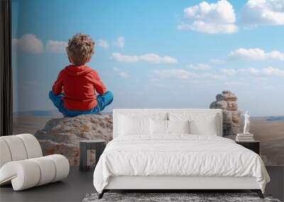 A young boy in a red hoodie sits meditatively on a large rock, gazing out over a breathtaking landscape with rock formations and a vibrant blue sky dotted with clouds. Wall mural