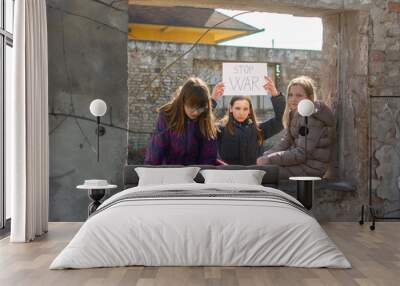 Three small girls in winter jackets standing on ruins of destroyed buildings in war zone, with Board No war, Stop war. Refugee children. The war in the Ukraine, combat actions. Wall mural