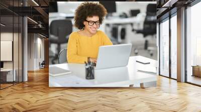 Beautiful smiling mixed race businesswoman dressed casual sitting in office and using laptop. Wall mural