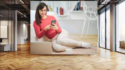 Young smiling woman sitting at home on the floor and using smart phone. Wall mural