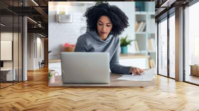 Afro business woman working with computer while consulting some invoices and documents in the kitchen at home. Wall mural