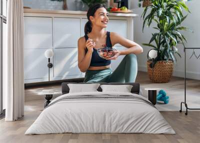 Athletic woman eating a healthy bowl of muesli with fruit sitting on floor in the kitchen at home Wall mural