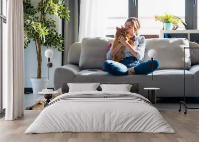 Attractive young woman kissing her little cute dog while sitting in couch with her dogs and cat in living room at home. Wall mural