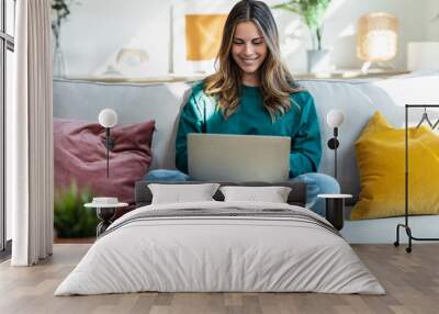 Beautiful kind woman working with laptop while sitting on couch in living room at home Wall mural