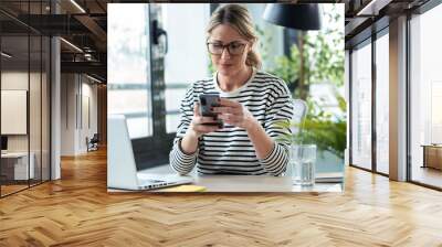 Beautiful mature business woman typing messages with her mobile phone while working with computer in the office at home. Wall mural