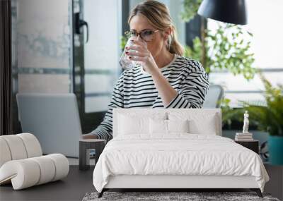 Beautiful mature business woman working with a laptop while drinking glass of water on a desk in the office at home. Wall mural