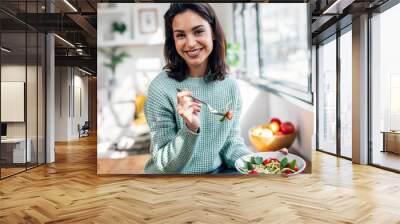 Beautiful smiling woman eating healthy salad while sitting on the table at home. Wall mural