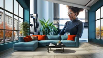 Beautiful young afro business woman working while making video call with computer sitting in the office. Wall mural